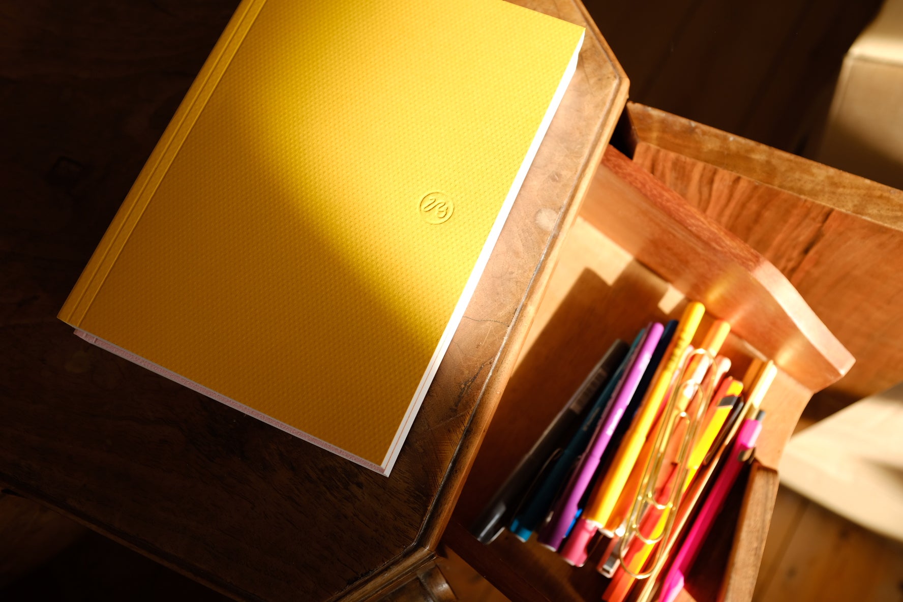 Yellow notebook on a wooden surface with a drawer open, showing pens and pencils.