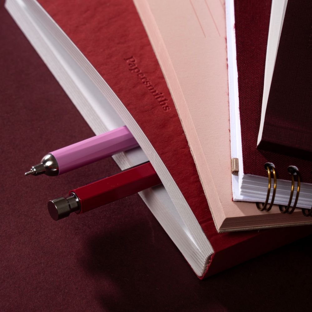 A close-up of a red hardcover notebook or journal with two pens tucked inside the cover, alongside other stacked books or notebooks on a dark burgundy surface.
